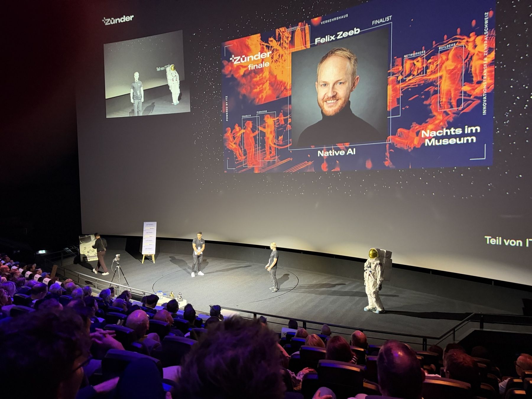 Felix Zeeb on stage at the Zünder finale in the Swiss Museum of Transport in Lucerne, his portrait on the screen with the words Finalist and Native AI, a full audience in front